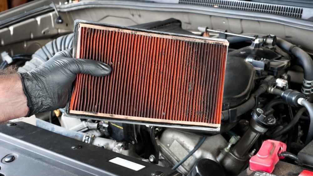 An auto mechanic wearing protective work gloves holds a dirty, clogged air filter over a car engine during general auto maintenance.